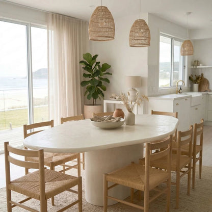 Dining room with a white table and wooden chairs, featuring a kitchen in the background.