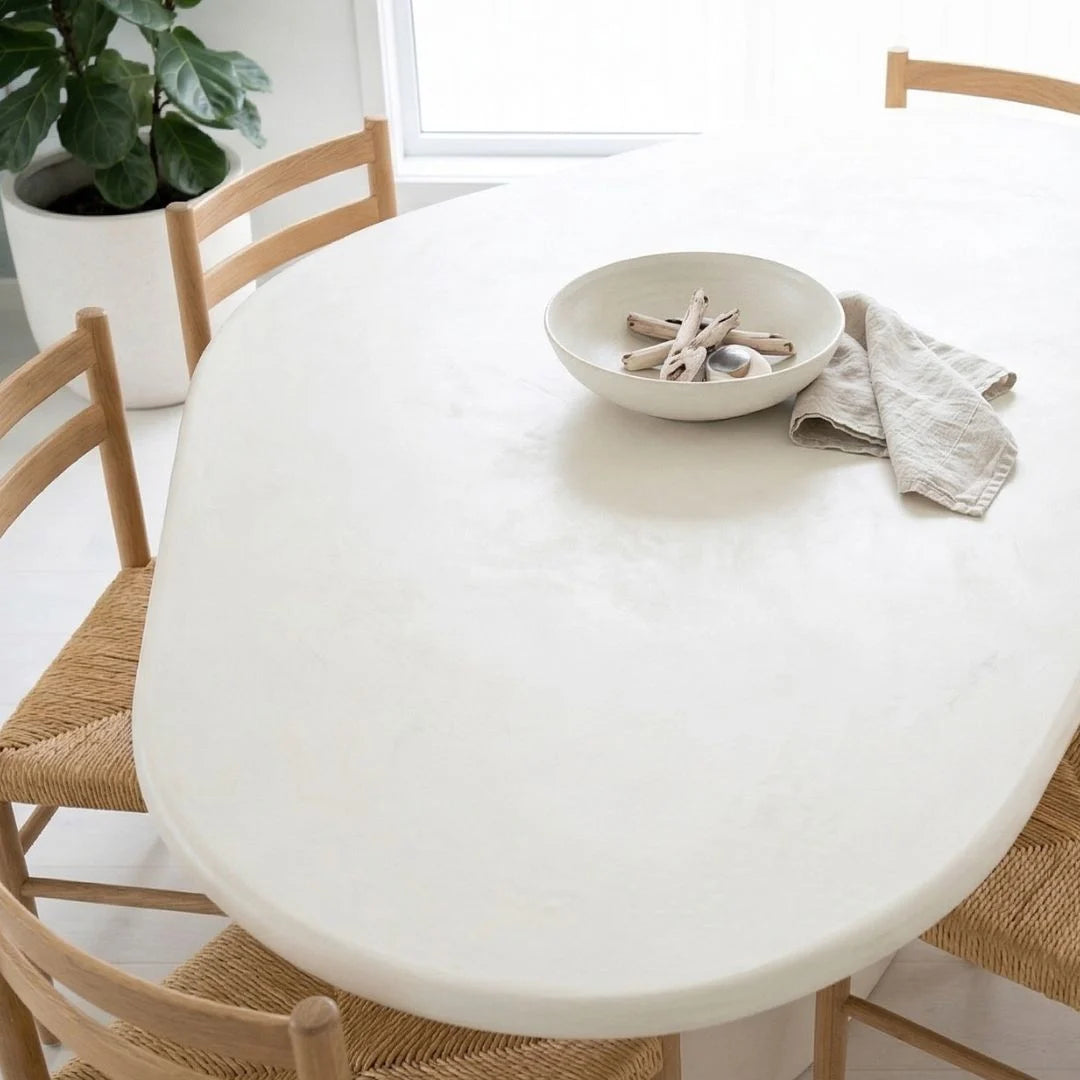 Round white table with a bowl and cloth on top, surrounded by wooden chairs in a bright room.