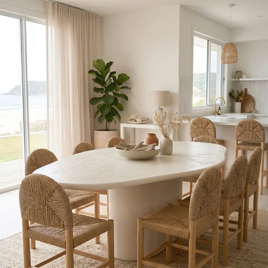 Dining room with a white table and wicker chairs, large windows, and a plant.