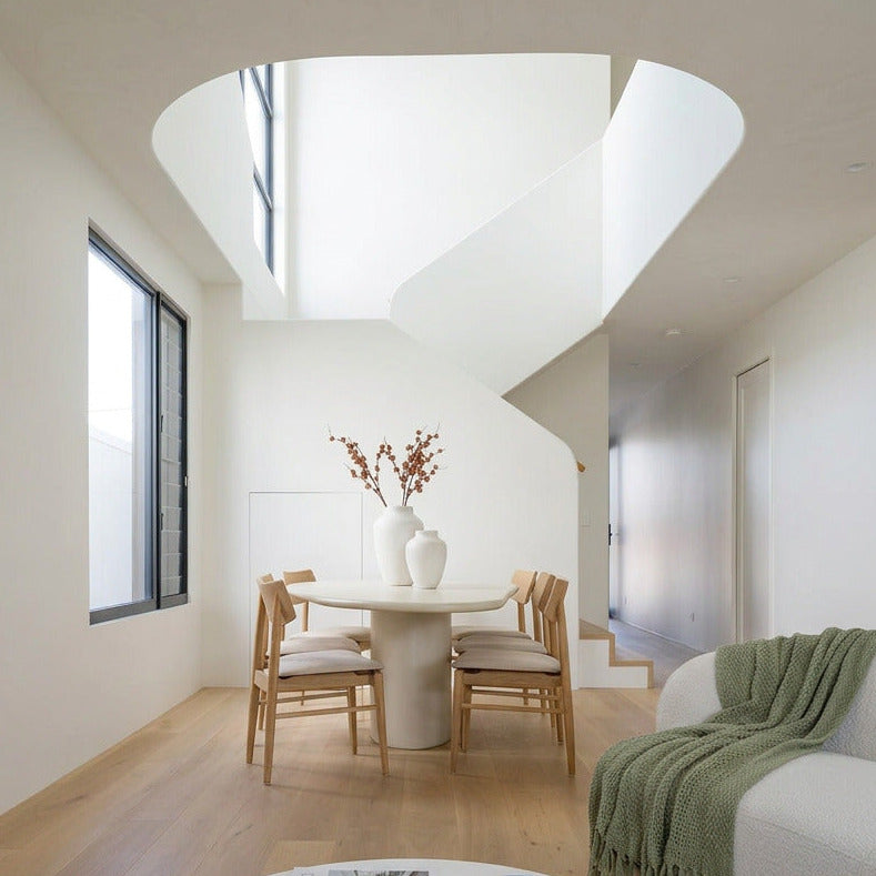 A bright, modern interior space with a round white coffee table in the foreground, a dining set with light wood chairs in the background, and a staircase opening above. The room has white walls, light wood flooring, and a green throw blanket on a sofa.