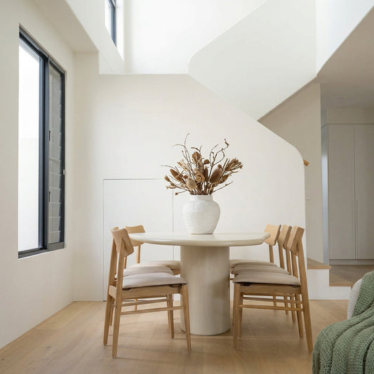 A minimalist dining set featuring a round off-white pedestal table and four light wood chairs with cream cushions, styled with a large white vase containing dried branches in the center. The room has white walls, a staircase in the background, and light wood flooring.