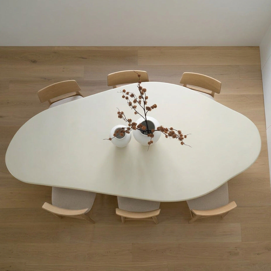 An overhead view of a modern organic-shaped white dining table surrounded by six light wood chairs, with a small floral centerpiece on a light wood floor.