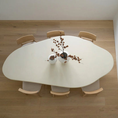 An overhead view of a modern organic-shaped white dining table surrounded by six light wood chairs, with a small floral centerpiece on a light wood floor.