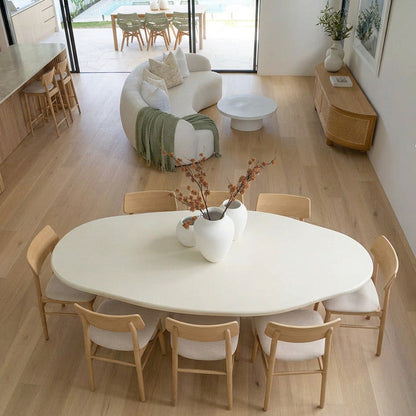 An overhead view of a modern dining room featuring a large, organic-shaped white dining table with light wood chairs, set in a bright room with light oak floors and a curved white sofa in the background.