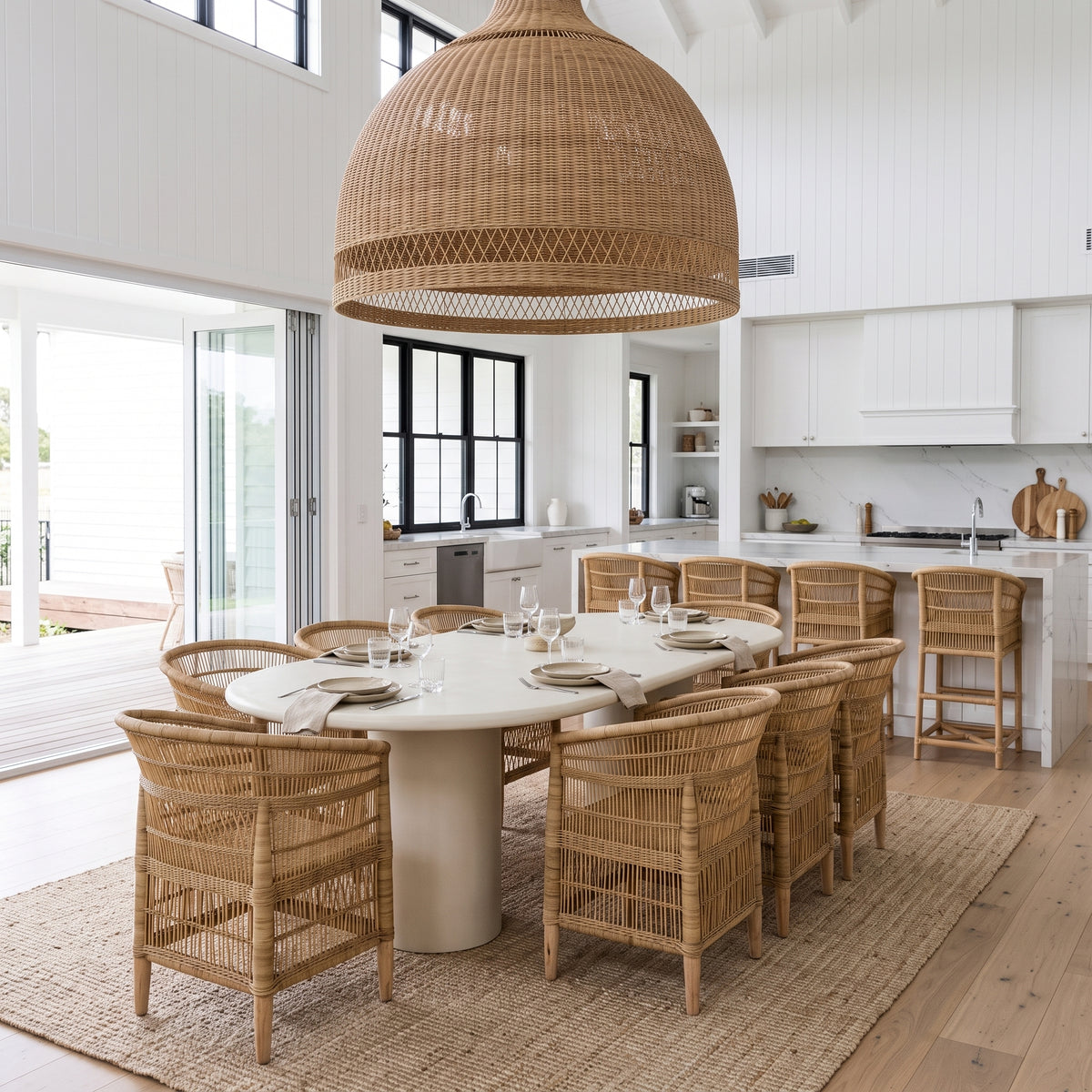 A large woven rattan dome pendant light hangs over a round dining table set with woven chairs in a bright, modern white dining room with a kitchen in the background.