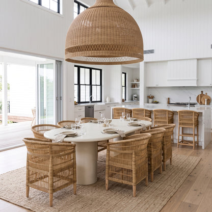 A large woven rattan dome pendant light hangs over a round dining table set with woven chairs in a bright, modern white dining room with a kitchen in the background.