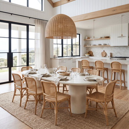A dining room scene with a round white dining table surrounded by woven natural-fiber chairs, set on a jute rug in a bright modern kitchen with large windows and a tall wicker pendant light overhead.
