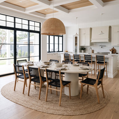 A dining room scene in a bright modern home with a round white dining table, eight wooden chairs with black woven seats, a large round natural jute rug, and a tall woven pendant light overhead.