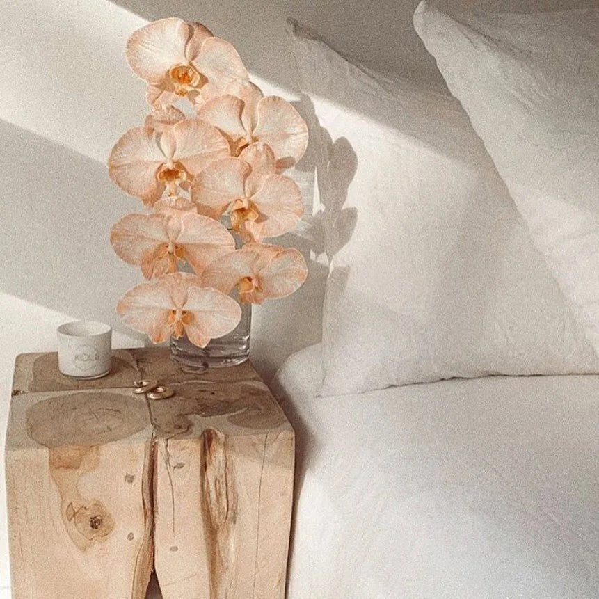 Vase with peach-colored orchids on a wooden side table next to a bed.