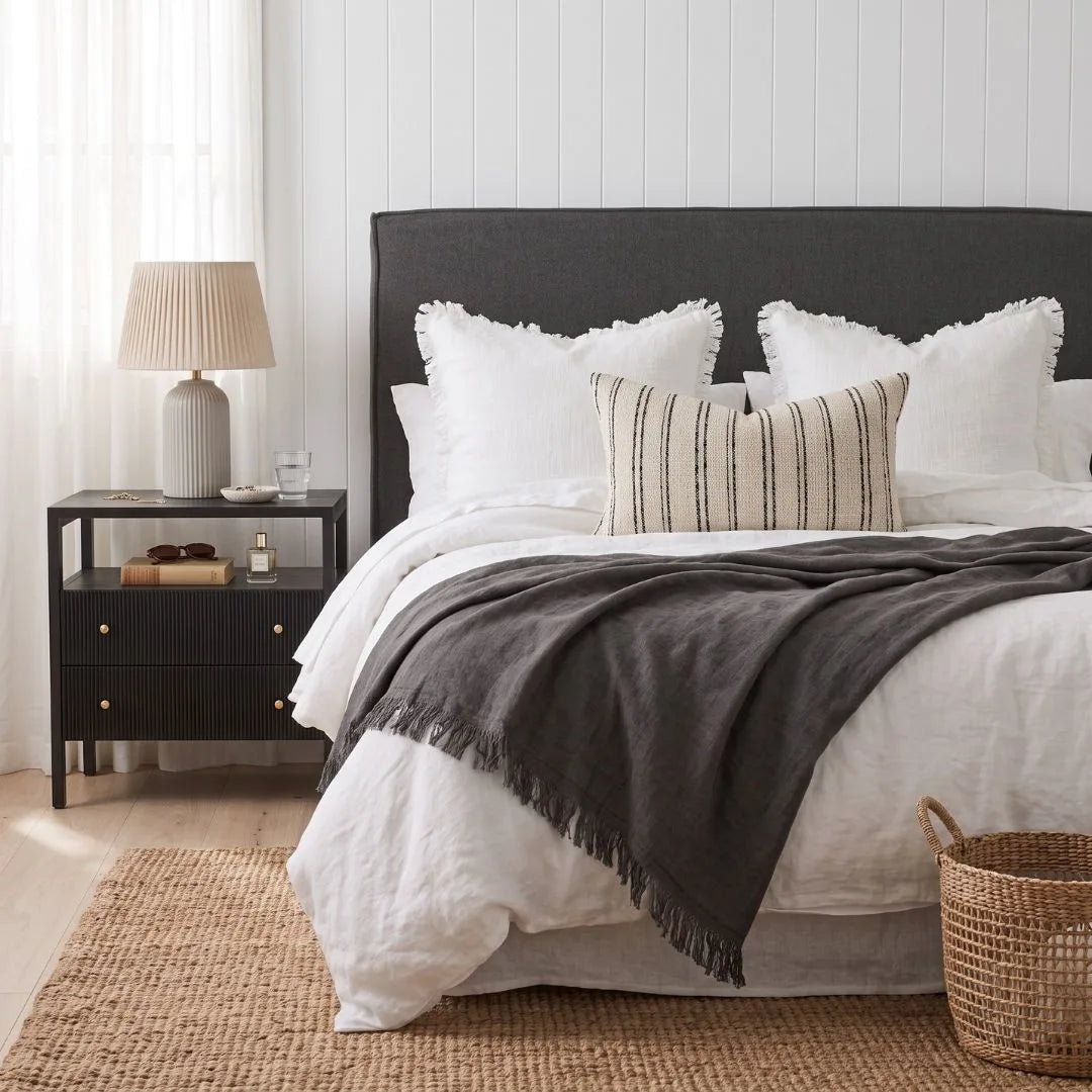 A styled bedroom scene showing a bed dressed in white bedding with a dark charcoal gray throw blanket and matching dark upholstered headboard. A striped beige accent pillow rests on the bed, and a black nightstand with a lamp is visible on the left.