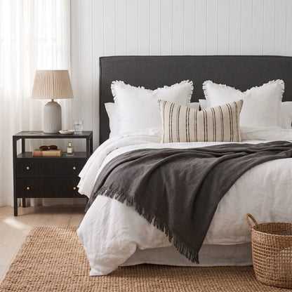 A styled bedroom scene showing a bed dressed in white bedding with a dark charcoal gray throw blanket and matching dark upholstered headboard. A striped beige accent pillow rests on the bed, and a black nightstand with a lamp is visible on the left.