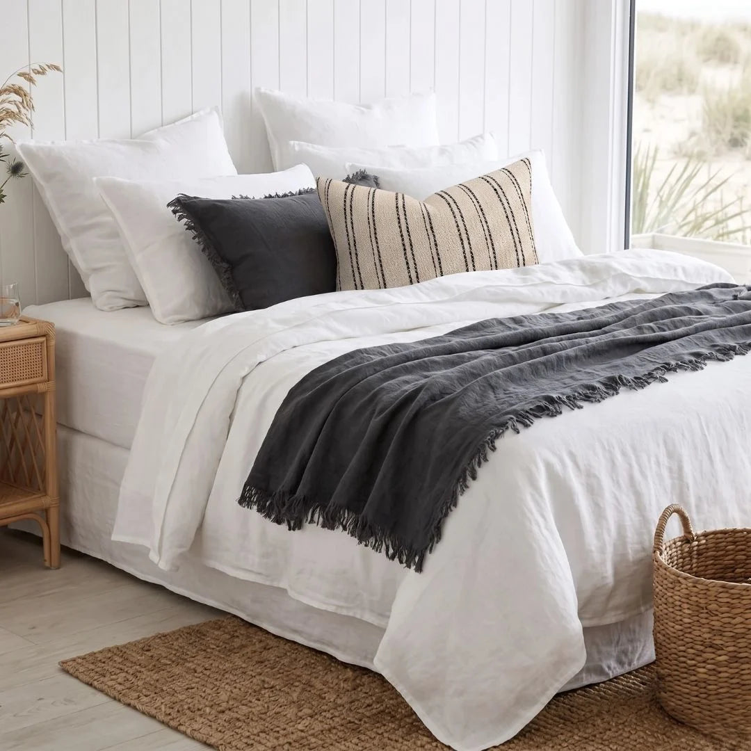 A neatly made bed with white bedding, a dark charcoal throw blanket draped across the foot, and two decorative pillows including one striped beige and black pillow, set in a bright bedroom with a window and woven rug.