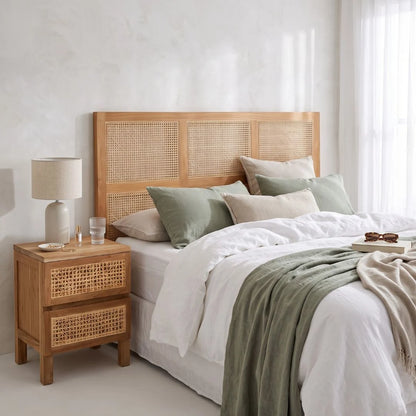A styled bedroom scene featuring a light natural wood bed frame with woven cane panels, dressed in white linens and sage green pillows, alongside a matching wooden nightstand with a lamp.
