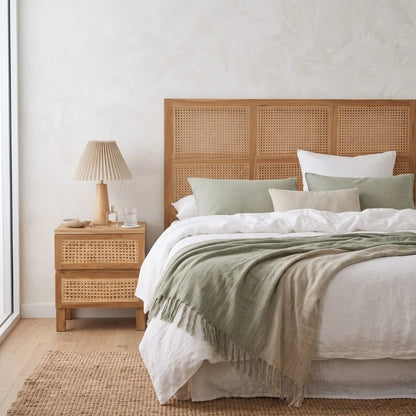 A styled bedroom scene featuring a bed with a light natural wood rattan headboard, white linens, sage green pillows and throw blanket, a matching rattan nightstand with a pleated beige lamp, and a textured beige rug against a white wall.