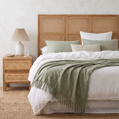 A styled bedroom scene featuring a bed with a light wood rattan headboard, white bedding, sage green pillows and throw blanket, a matching wood nightstand with a pleated white lamp, and a woven rug on a light wood floor.