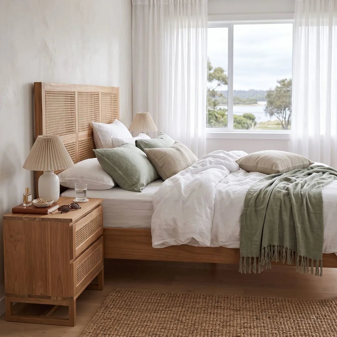 A styled bedroom scene featuring a light natural wood bed with a woven cane headboard, dressed in white linens and sage green pillows and a fringed throw blanket, next to a matching wooden nightstand with a lamp.
