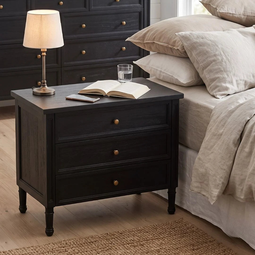 A dark wood nightstand with three drawers and brass knobs, styled in a bedroom next to a bed with beige linens. A table lamp, an open book, and a glass of water sit on top.