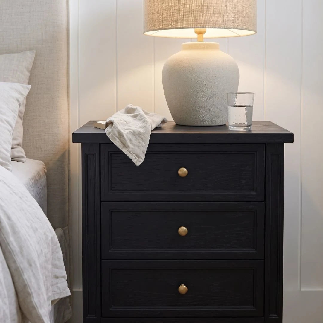 A black three-drawer nightstand with round gold knobs, styled next to a bed with white bedding. On top of the nightstand sits a beige ceramic table lamp with a matching lampshade and a clear glass of water.