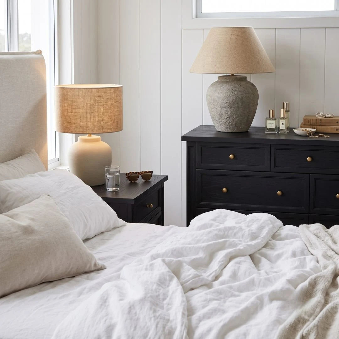 A styled bedroom scene with a bed in the foreground, a black nightstand beside it, and a matching black dresser holding a large textured beige table lamp. The room has white paneled walls, a window, and neutral bedding.