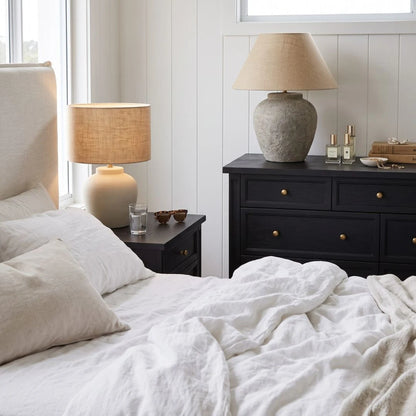 A styled bedroom scene with a bed in the foreground, a black nightstand beside it, and a matching black dresser holding a large textured beige table lamp. The room has white paneled walls, a window, and neutral bedding.