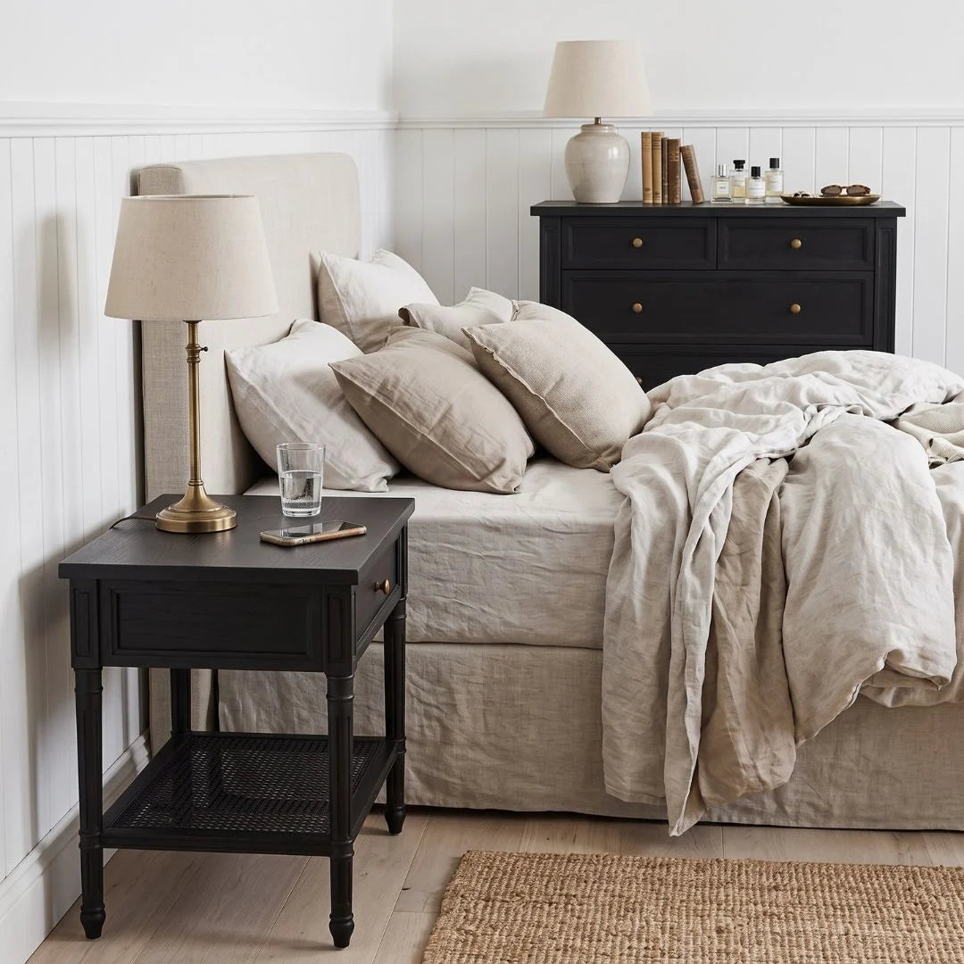 A styled bedroom scene featuring a black wooden nightstand with a table lamp and glass on top, next to a bed with beige linens and pillows.