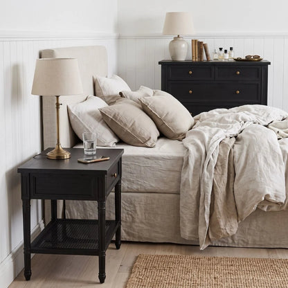 A styled bedroom scene featuring a black wooden nightstand with a table lamp and glass on top, next to a bed with beige linens and pillows.