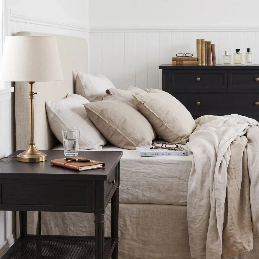 A styled bedroom scene featuring a black wooden nightstand with a table lamp and glass on top, next to a bed with beige linens and pillows.