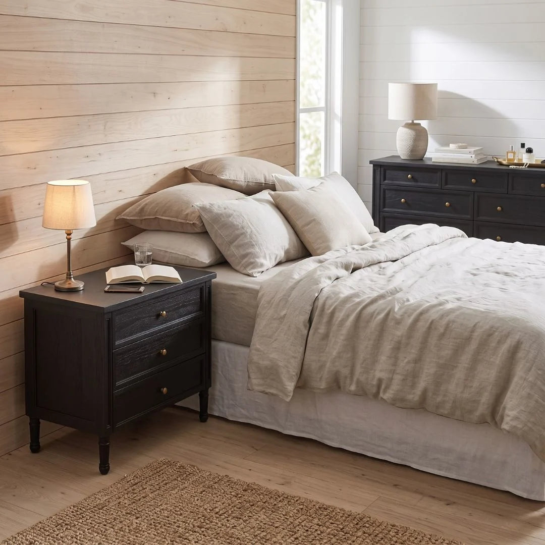 A styled bedroom scene showing a bed dressed in light beige linens, with a dark wood nightstand on the left holding a lit table lamp and an open book, and a matching dark wood dresser on the right against a window.