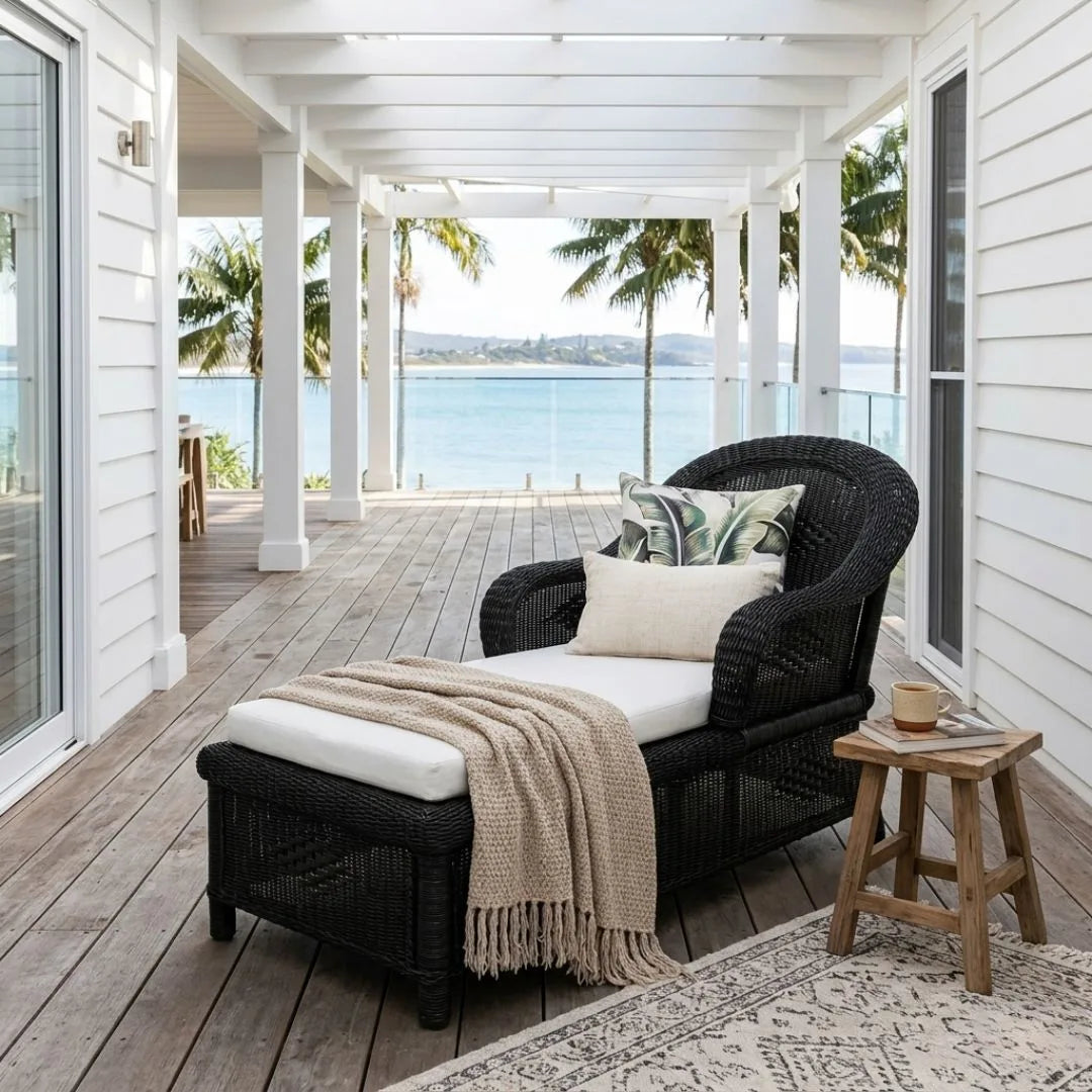 Black wicker chair with white cushion and beige blanket on a wooden deck with ocean view
