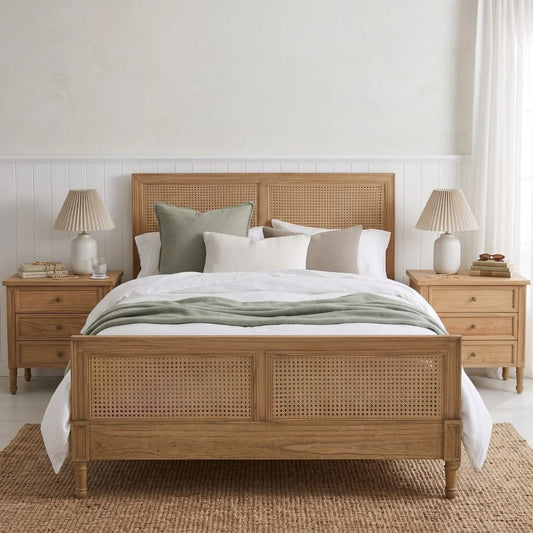 A light natural wood bed frame with woven cane panels, styled with white and sage green bedding, flanked by matching wooden nightstands with lamps.