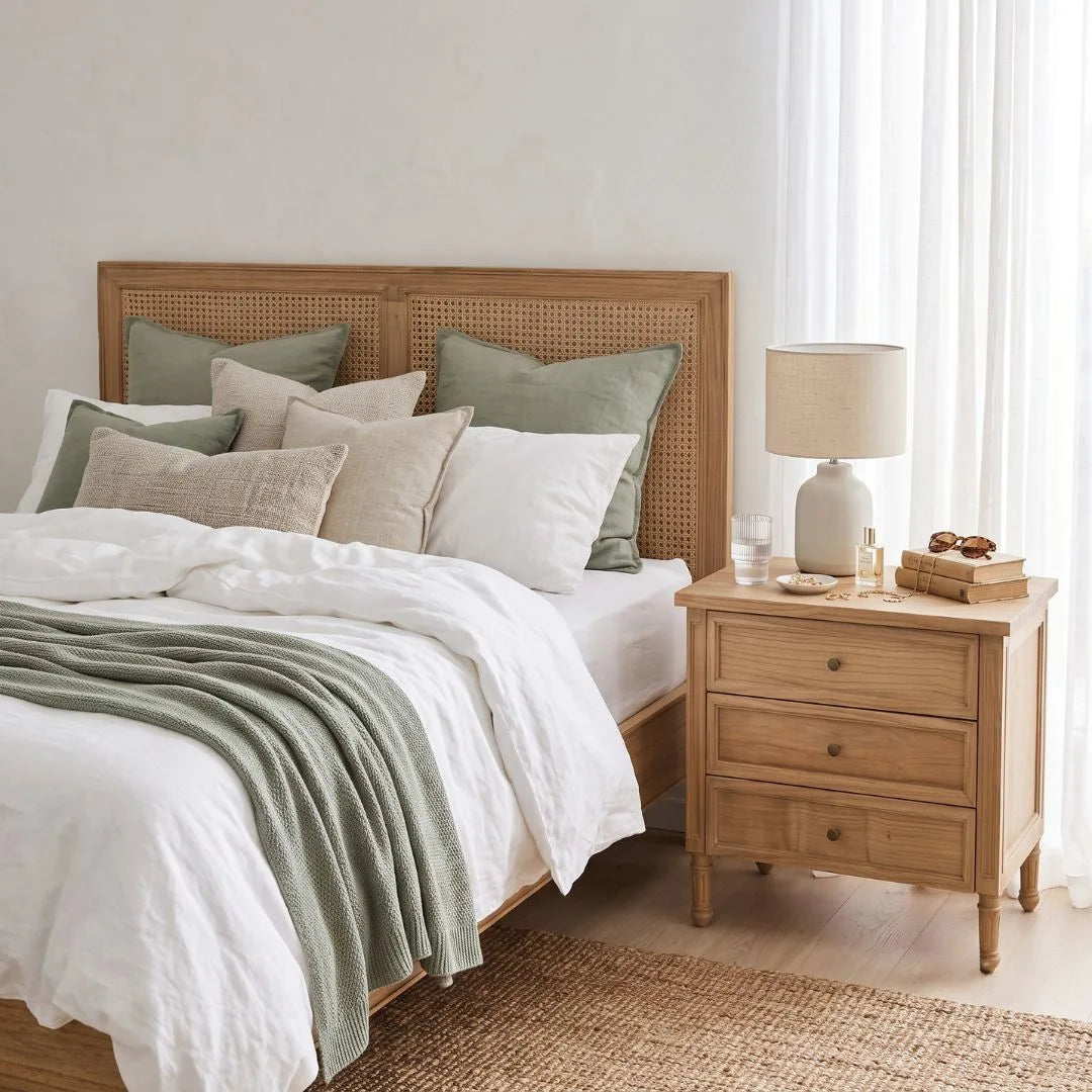 A styled bedroom scene showing a light natural wood bed frame with a woven cane headboard, dressed in white linens and sage green pillows and a throw blanket, next to a matching wood three-drawer nightstand with a table lamp.