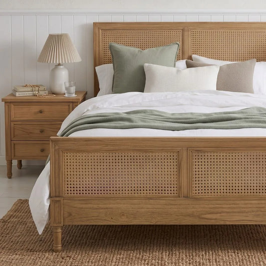 A light oak wood bed frame with woven cane panels, styled with white and sage green bedding, set in a bright bedroom next to a matching wooden nightstand.