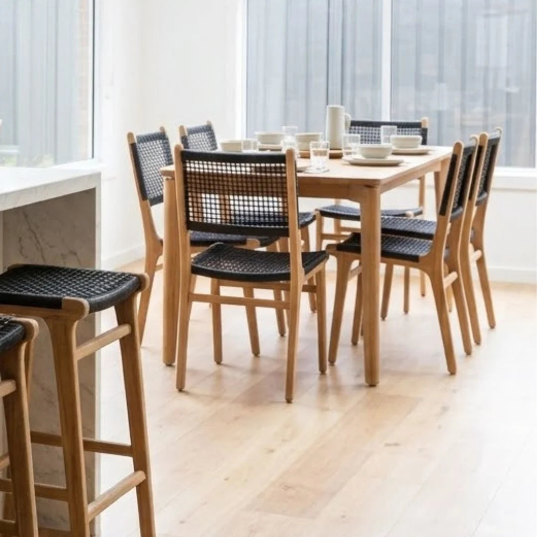 A light oak dining table with six matching chairs featuring black woven seats and backs, set in a bright, modern dining room with light wood floors and large windows.