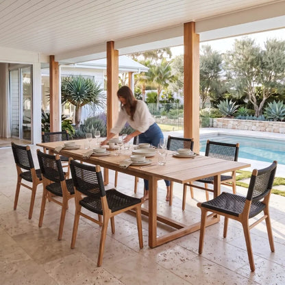 A woman sets a table with white dishes on a large wooden dining table with black woven chairs, situated on a covered patio overlooking a swimming pool and landscaped garden.
