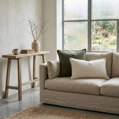 Beige sofa with green and white pillows in a room with a wooden side table and large window.