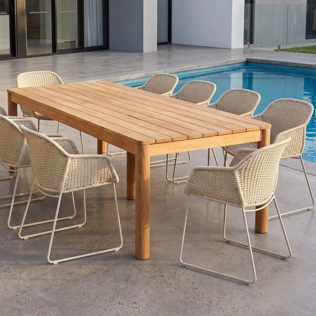 A rectangular outdoor dining table made of warm-toned natural wood slats, surrounded by eight woven wicker chairs with white metal frames, set on a concrete patio next to a swimming pool.