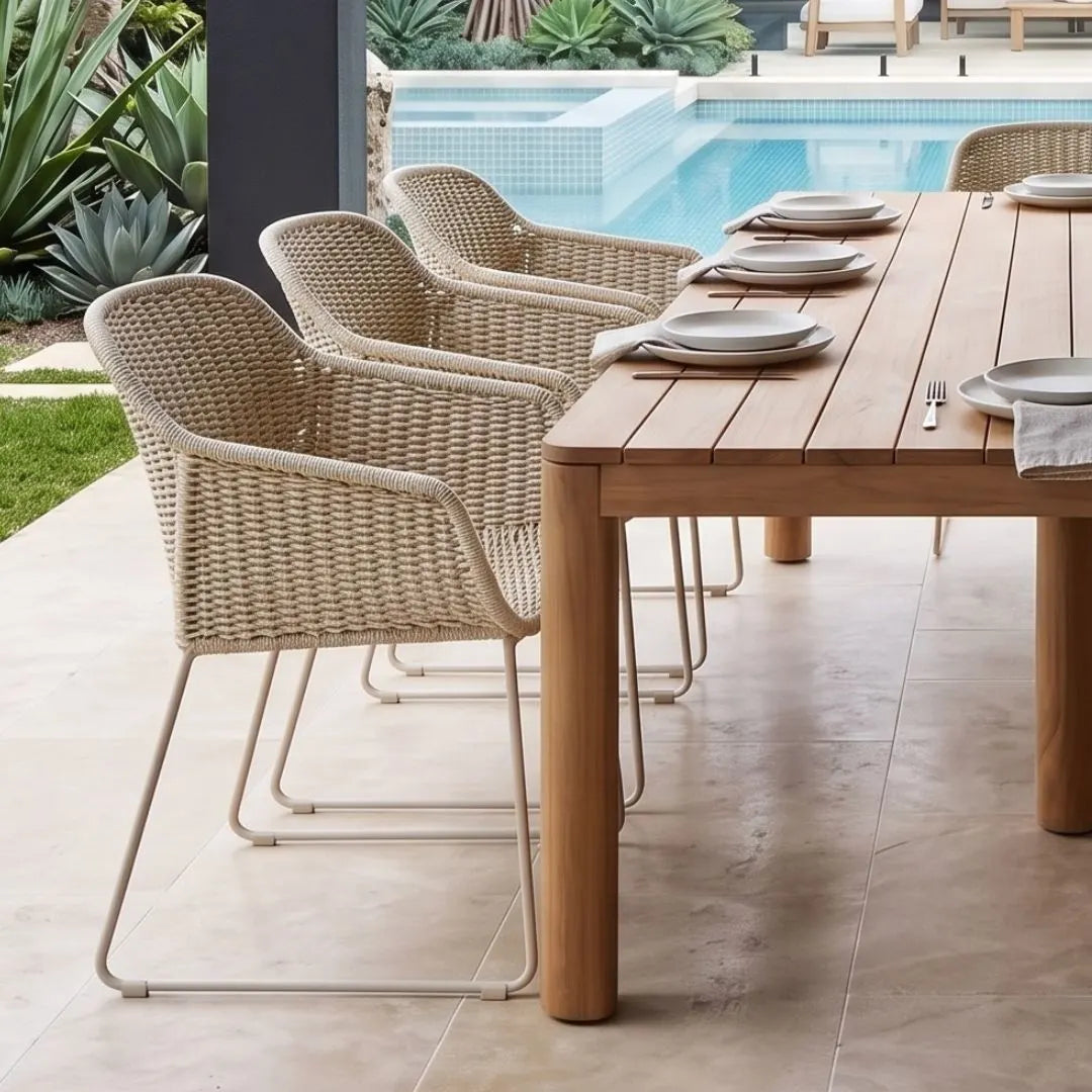 Outdoor dining set featuring a rectangular wooden table and four woven wicker armchairs with slim metal legs, arranged on a tiled patio beside a swimming pool.