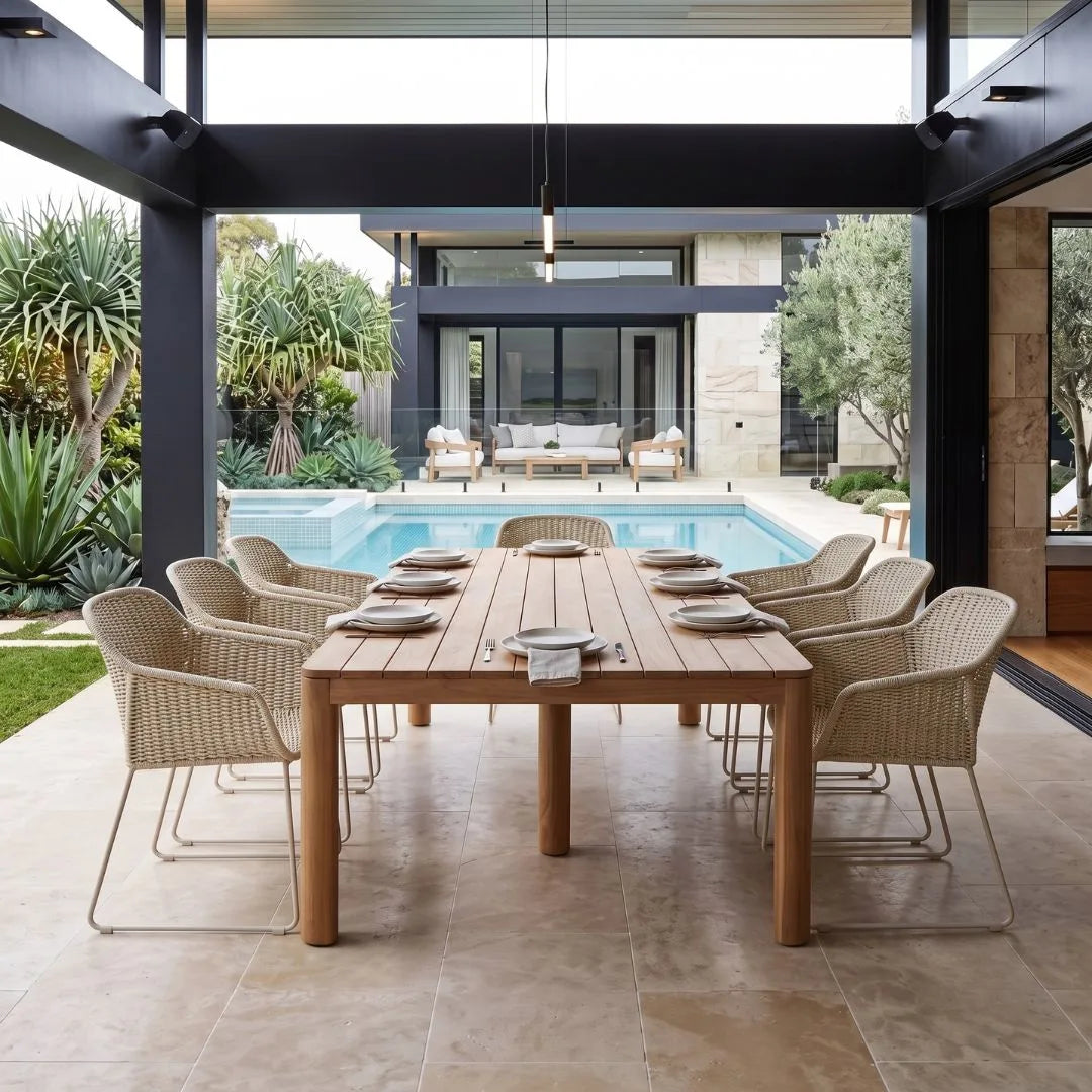 A modern outdoor dining setup featuring a rectangular wooden dining table surrounded by eight woven wicker armchairs with slim metal legs, set under a covered patio with a swimming pool and contemporary house in the background.