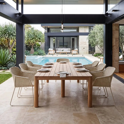 A modern outdoor dining setup featuring a rectangular wooden dining table surrounded by eight woven wicker armchairs with slim metal legs, set under a covered patio with a swimming pool and contemporary house in the background.