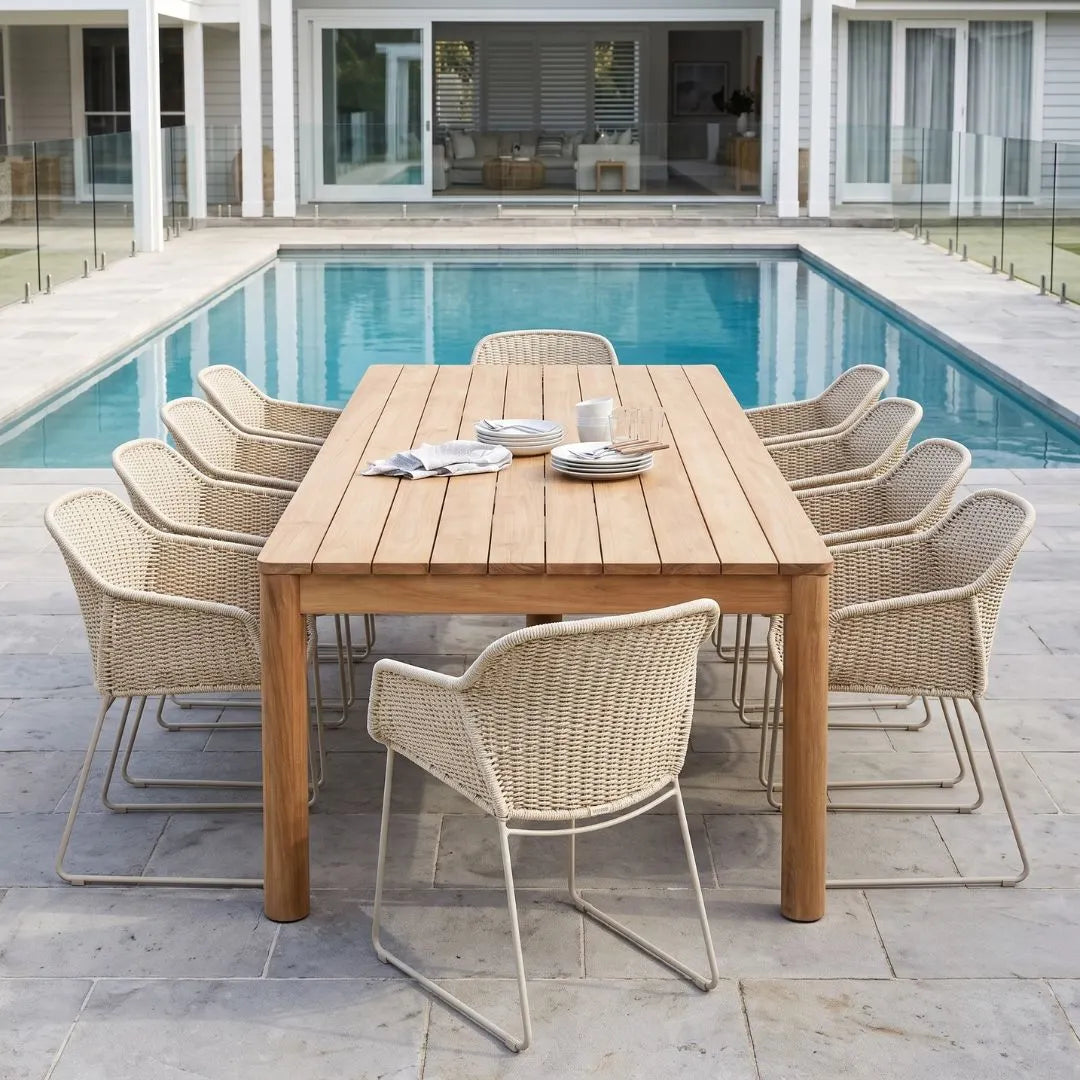 An outdoor dining set consisting of a rectangular light oak wood table and eight woven wicker armchairs with slim metal legs, arranged on a stone patio next to a swimming pool.