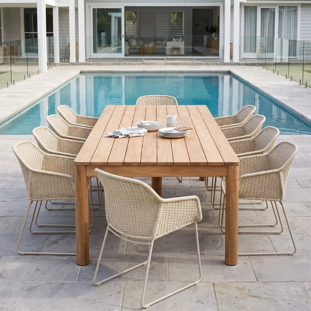 Outdoor dining set with a rectangular light wood table and eight woven wicker chairs with metal legs, arranged on a stone patio with a swimming pool and modern house in the background.