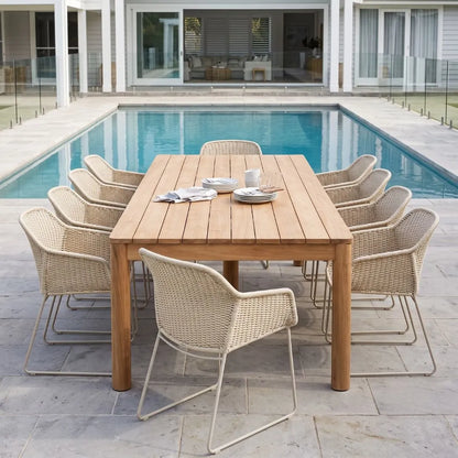 Outdoor dining set with a rectangular light wood table and eight woven wicker chairs with metal legs, arranged on a stone patio with a swimming pool and modern house in the background.