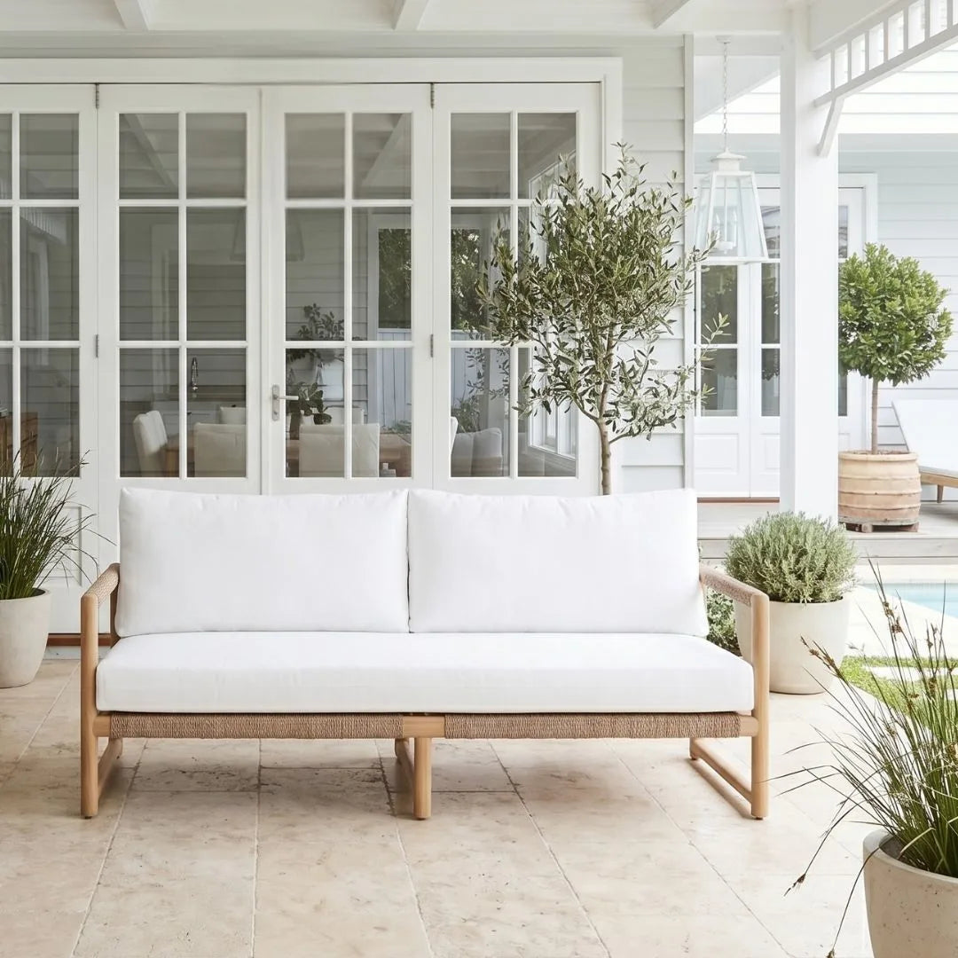 White cushioned outdoor sofa with wooden frame in a bright, sunlit room with large windows and plants.
