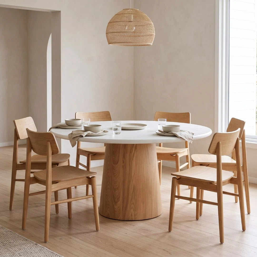 A round dining table with a white top and a natural wood pedestal base, surrounded by six matching wooden chairs in a bright, modern dining room.