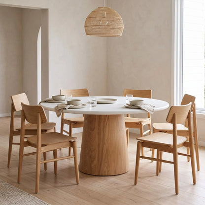 A round dining table with a white top and a natural wood pedestal base, surrounded by six matching wooden chairs in a bright, modern dining room.