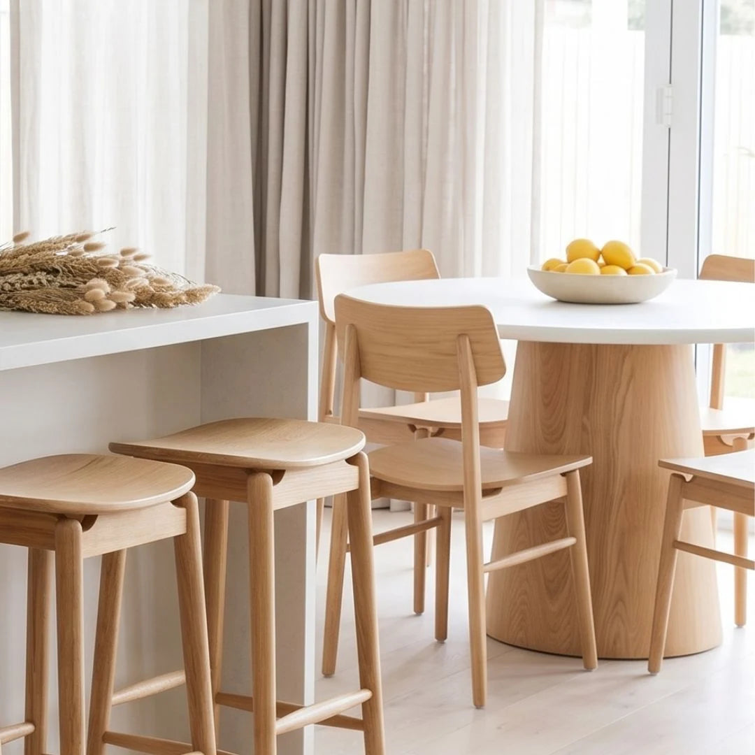 A modern dining set in a bright, minimalist room, featuring a round white table with a tapered light wood base, matching light wood chairs, and two matching bar stools. A white bowl of lemons and a decorative wheat arrangement sit on the table and counter.