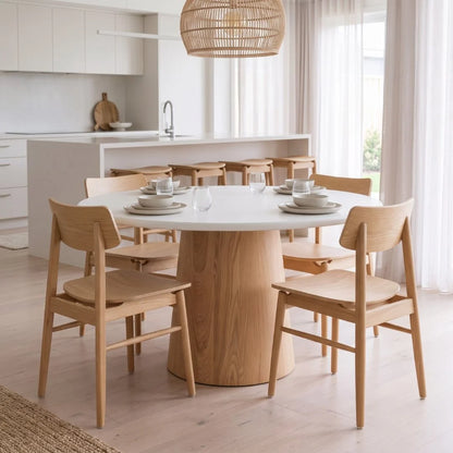 A round dining table with a thick natural wood pedestal base and a white top, surrounded by six light natural wood chairs with curved backs, set in a bright, modern kitchen.