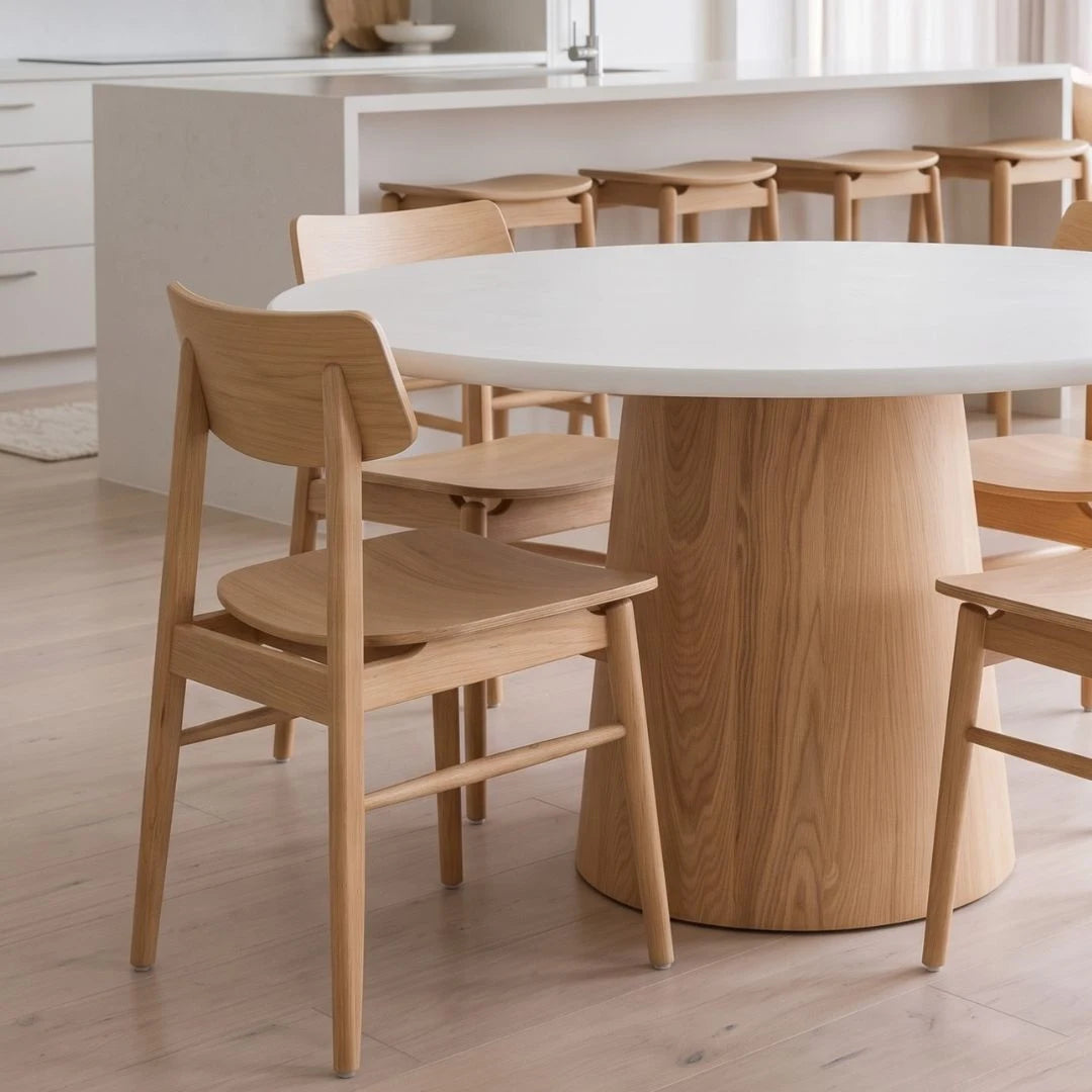 A light natural wood dining table with a thick cylindrical pedestal base and matching wooden chairs, set in a bright, modern kitchen interior.