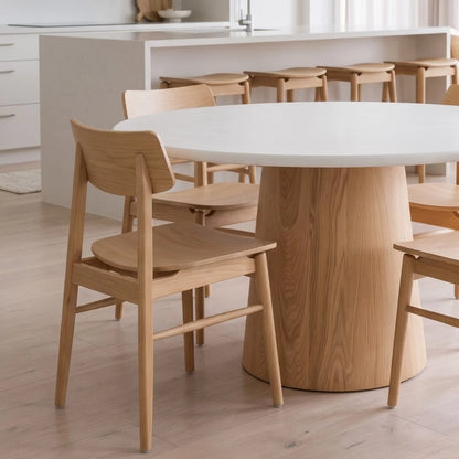 A light natural wood dining table with a thick cylindrical pedestal base and matching wooden chairs, set in a bright, modern kitchen interior.