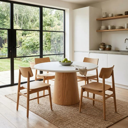 A round dining table with a white top and a natural wood pedestal base, surrounded by four matching wooden chairs in a bright, modern kitchen-dining room with large windows and outdoor greenery.